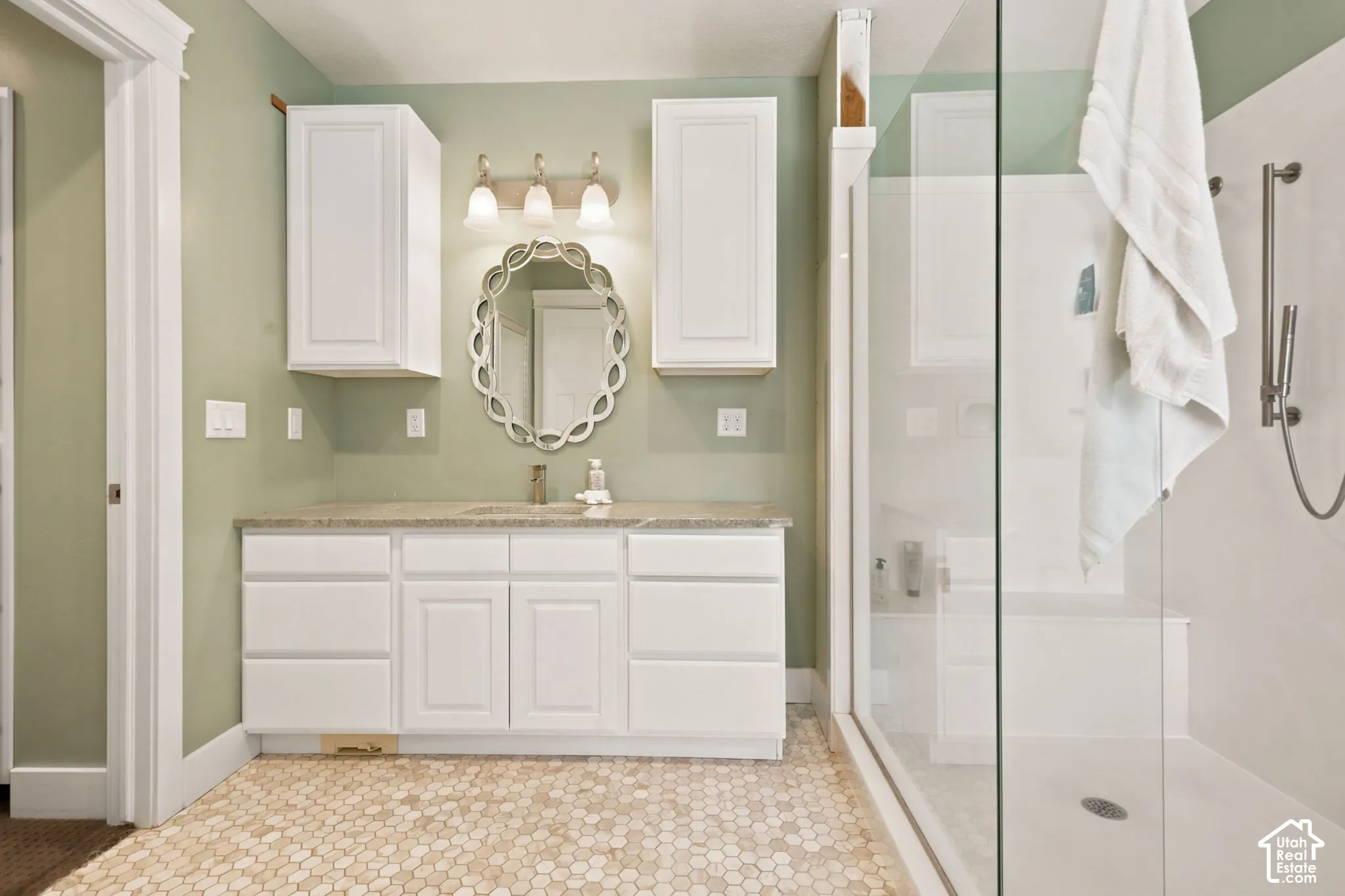 Bathroom featuring a clear glass shower enclosure with a built-in bench, hexagon tile flooring, a single vanity with stone-finish countertop, white cabinetry, and a decorative wall.   Digitally staged.
