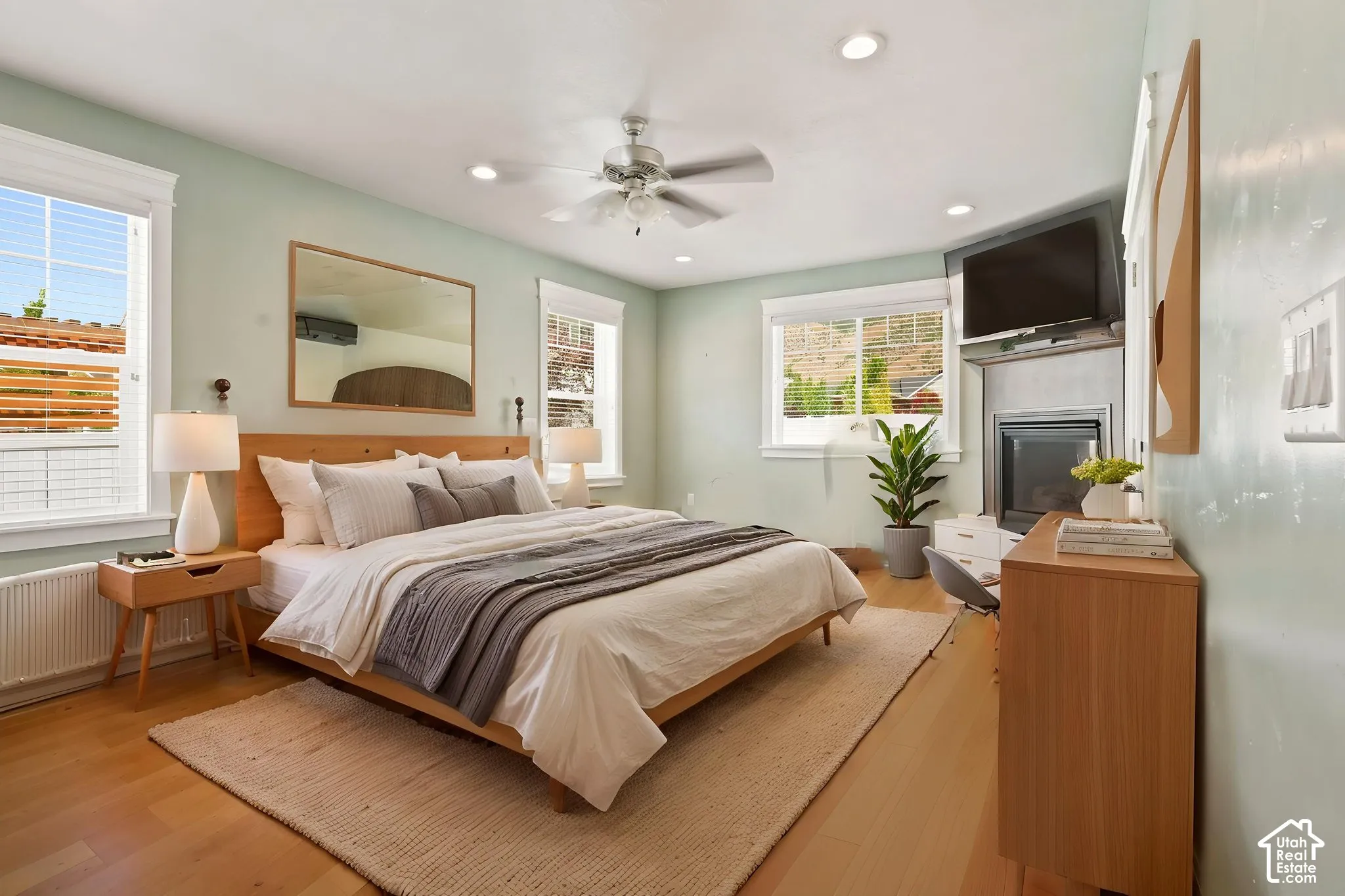 Hardwood flooring and light green walls frame a bedroom featuring a built-in fireplace with a television   Digitally staged.