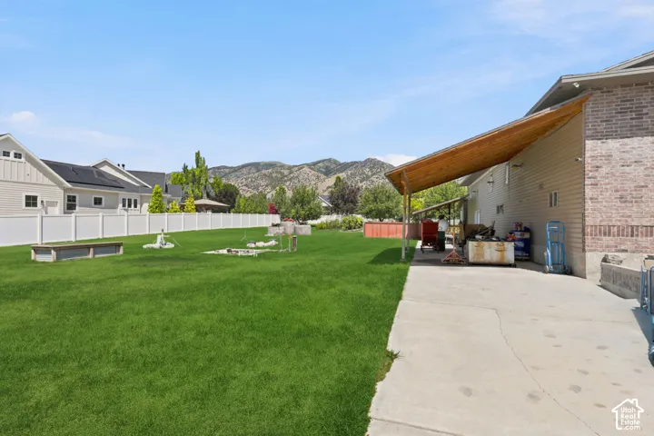 Expansive grassy yard with mountain backdrop, featuring a white privacy fence, concrete patio, and a covered wood-beam awning