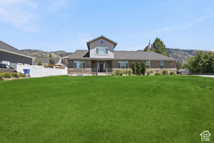 Expansive front lawn with a brick and siding exterior, a white picket fence, and a mountain backdrop  Digitally staged.