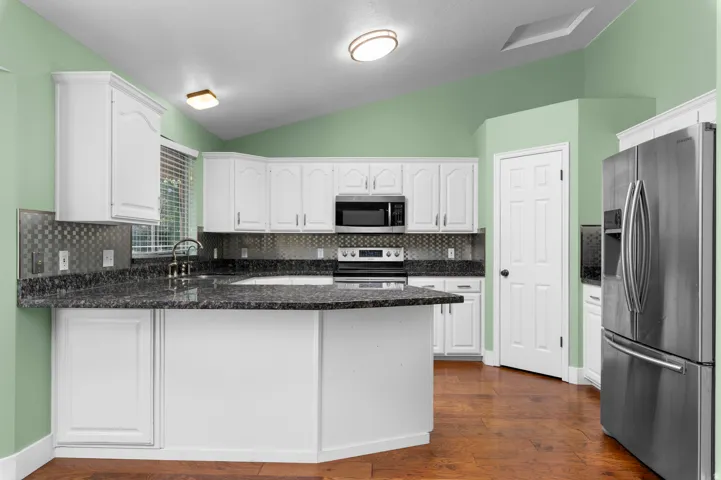 White cabinetry, stainless steel appliances, dark stone countertops, wood-finish flooring, and a tiled backsplash
