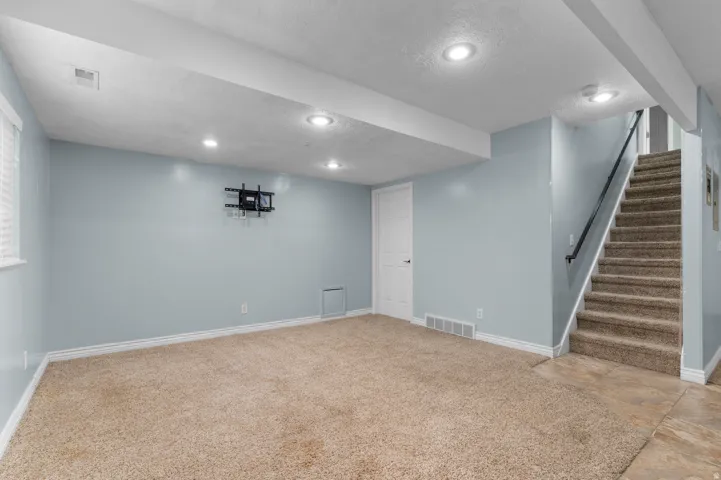 Expansive finished area with recessed lighting, carpet flooring, a white paneled door, and a staircase with a black handrail