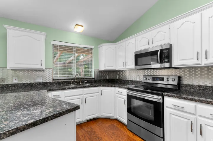 White cabinetry with stainless steel appliances, dark stone countertops, mosaic tile backsplash, and wood-finish flooring