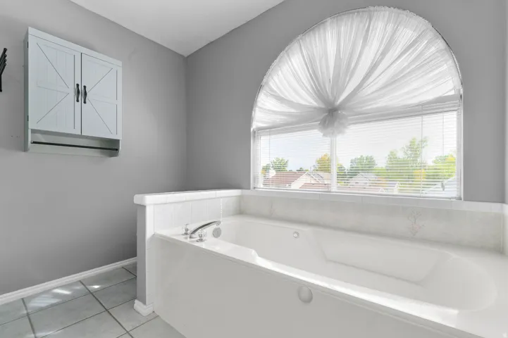 Primary Bedroom spa-inspired bathroom featuring a shower and separate jetted tub with integrated deck, arched transom window with sheer drapery, ceramic tile flooring, soft gray wall finishes, and wall-mounted cabinetry with geometric paneling detail