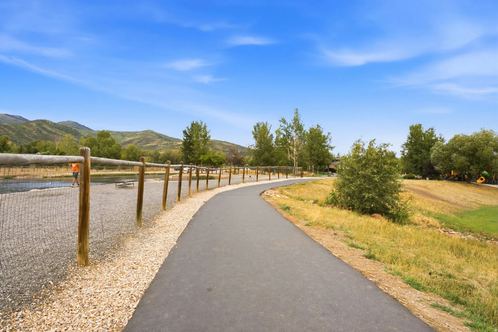 Paved pathway with a wood post and wire fence, bordered by a gravel and grass area, with distant mountains