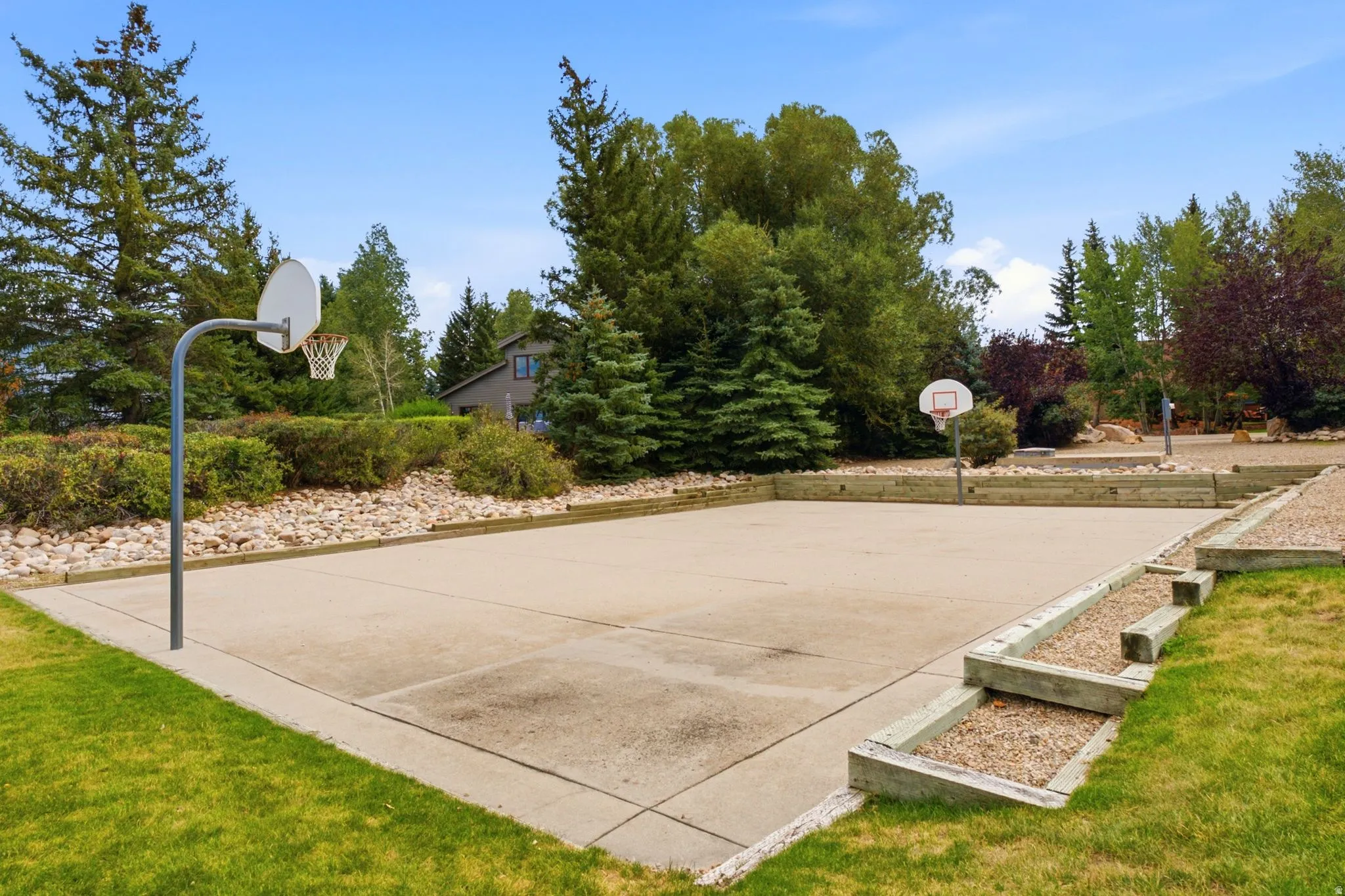 Paved sports court featuring two basketball hoops, surrounded by landscaped terraced beds and mature evergreen trees