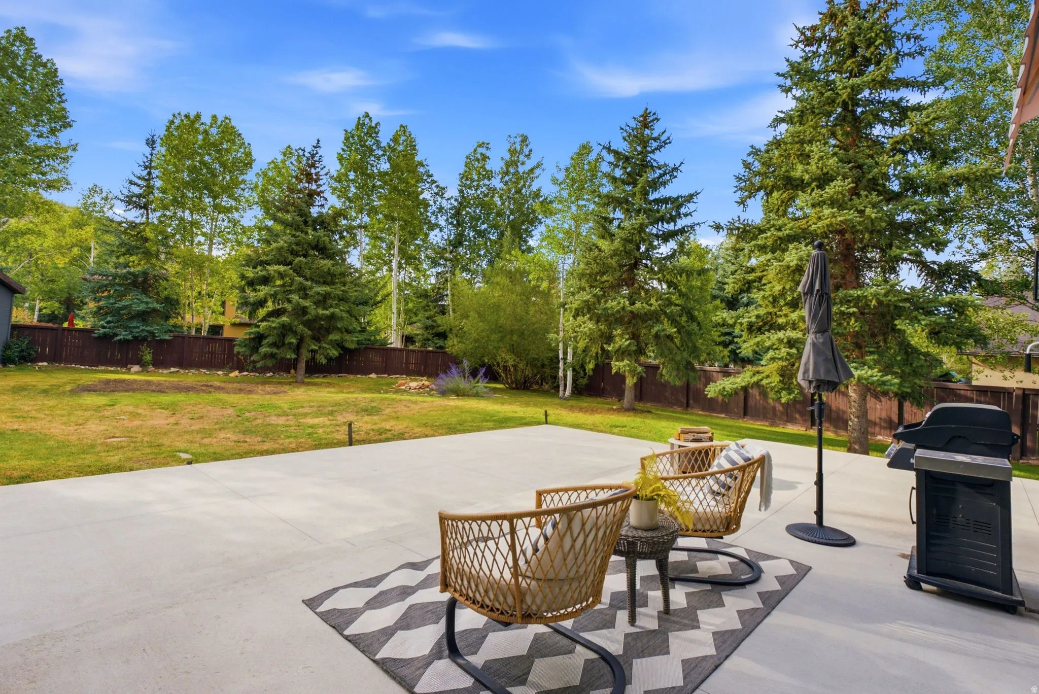 Expansive concrete patio with a spacious grassy area, perimeter fencing, and mature evergreen trees