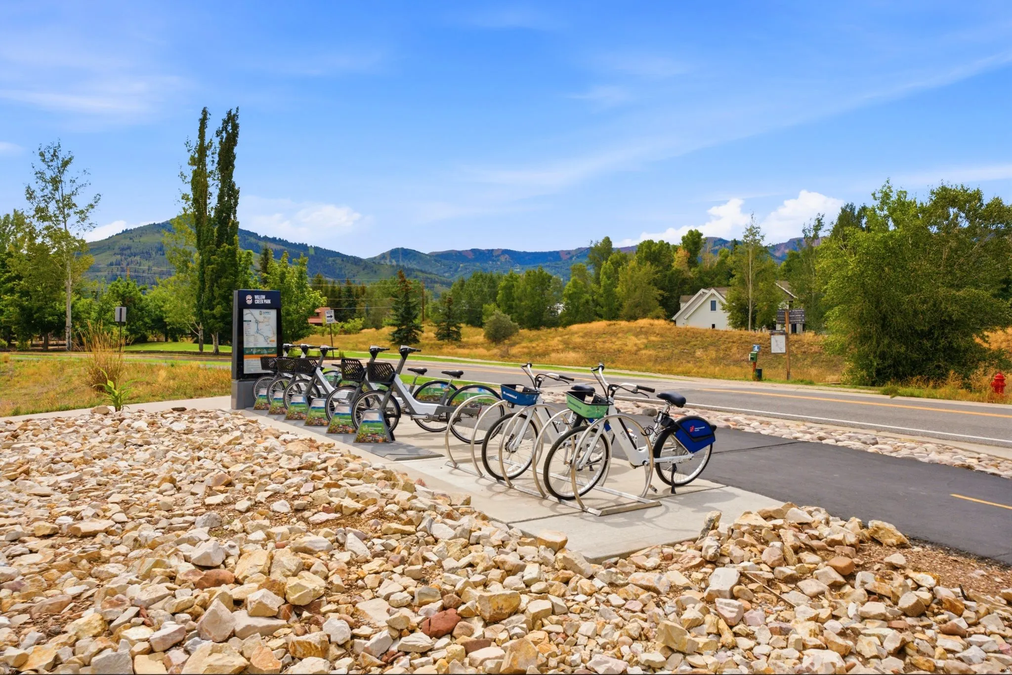 Paved pathway, bike share station, natural rock landscaping, distant mountain scenery, and mature trees
