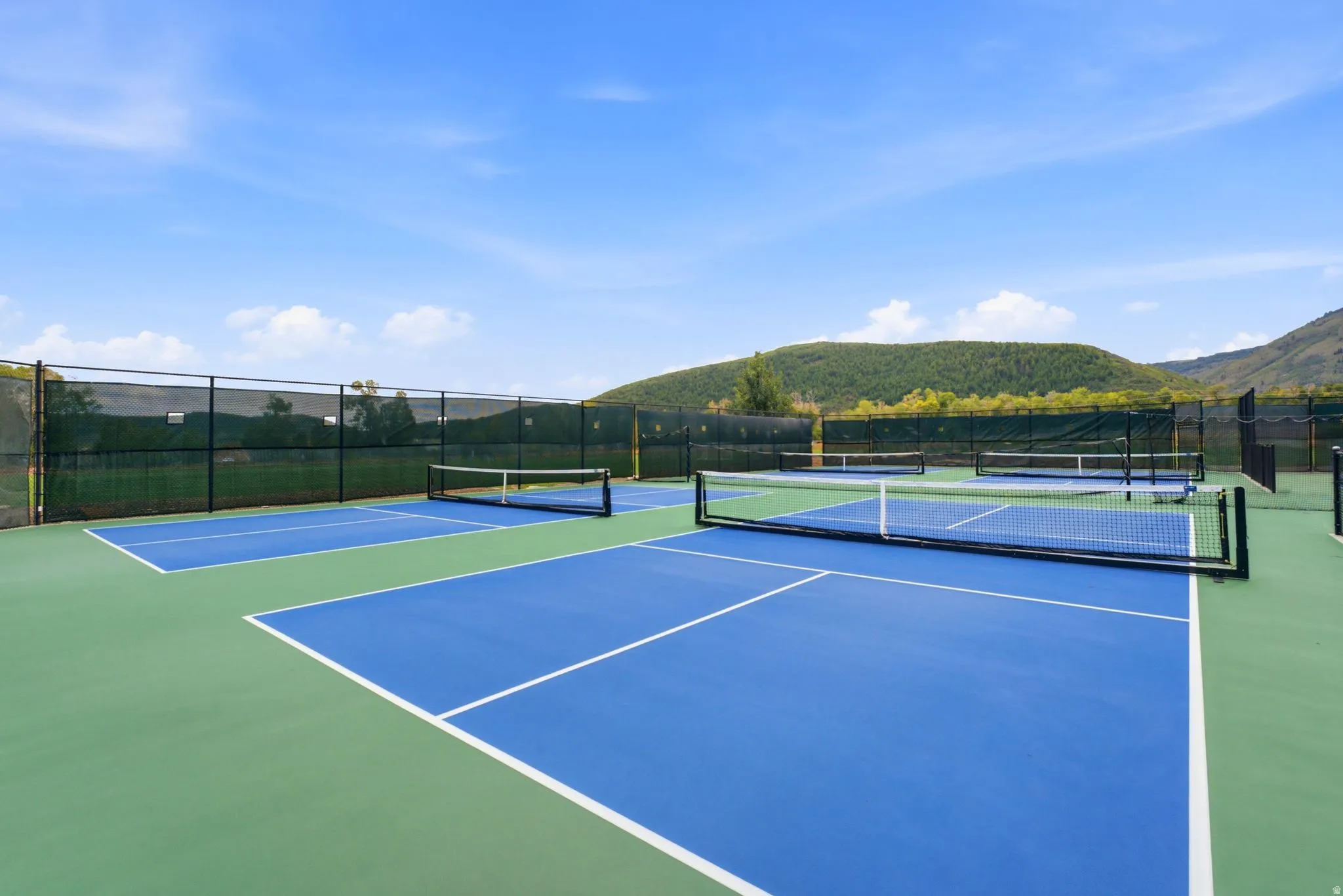 Multiple sport courts with blue and green surfacing, surrounded by black fencing, set against a backdrop of distant hills