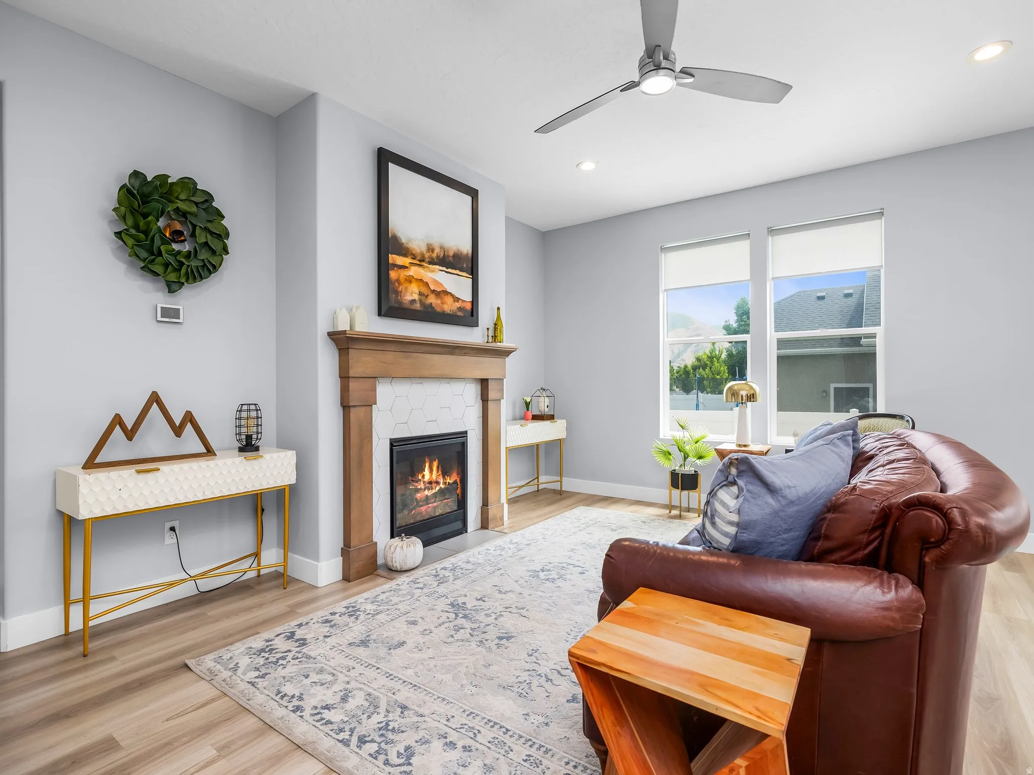 Living area featuring a gas fireplace with a wood mantle and hexagonal tile surround, light wood-finish flooring, two large windows, recessed lighting, and a ceiling fan