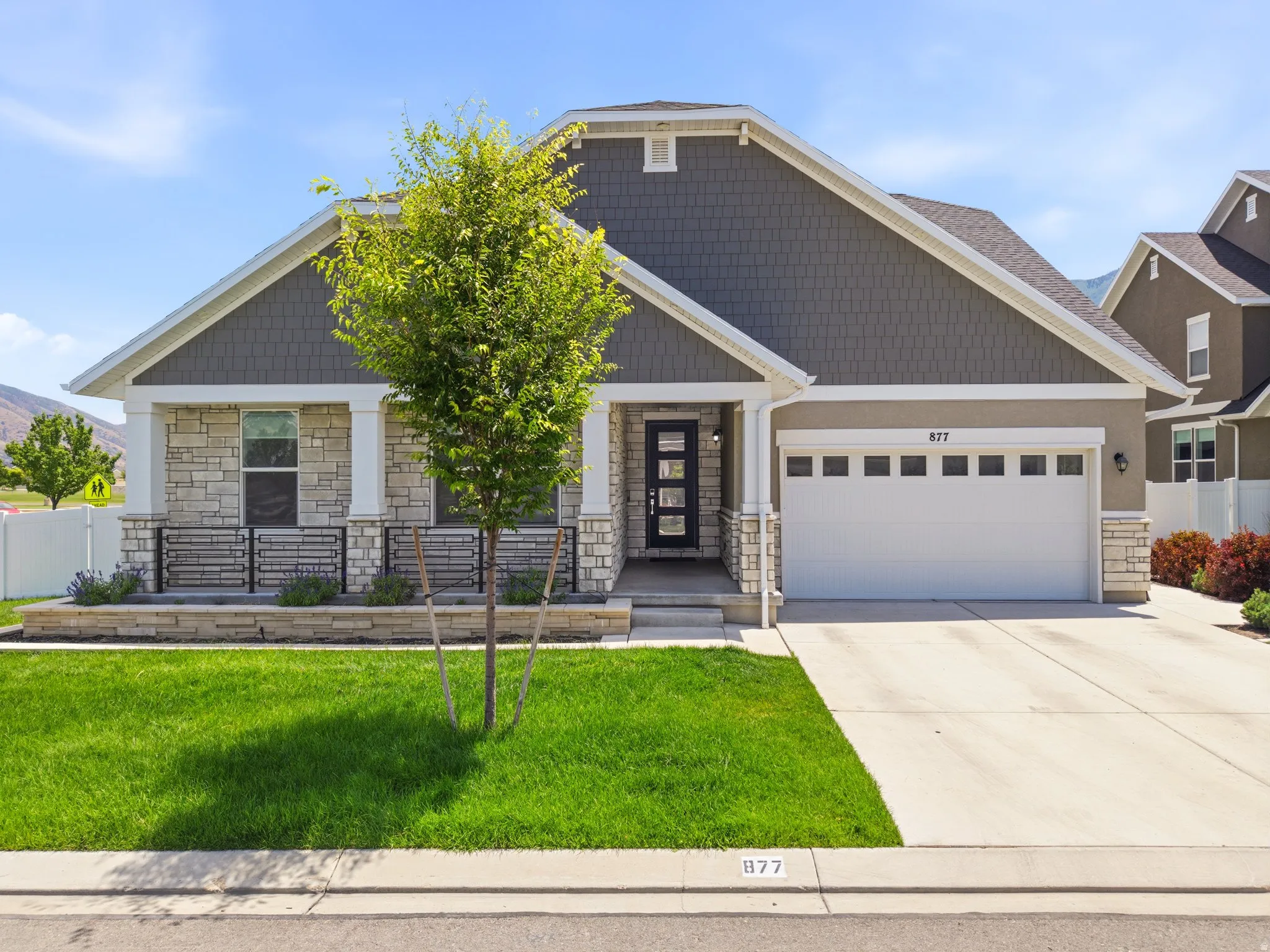 Contemporary facade featuring stacked stone accents, horizontal siding, a prominent front entry with a multi-pane door, an attached garage with upper windows, and a landscaped front yard with a young tree