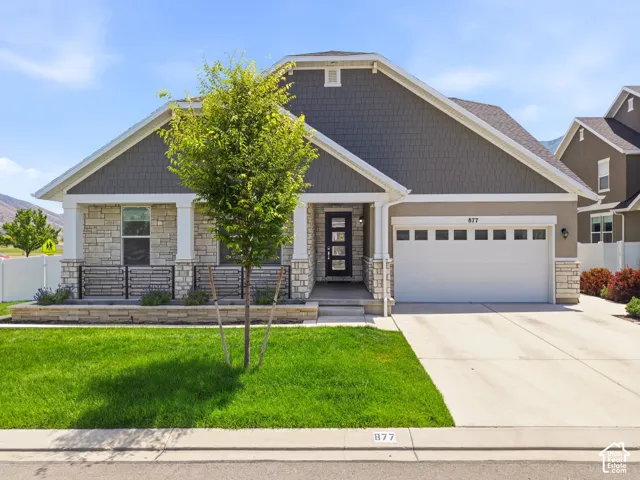 Contemporary facade featuring stacked stone accents, horizontal siding, a prominent front entry with a multi-pane door, an attached garage with upper windows, and a landscaped front yard with a young tree