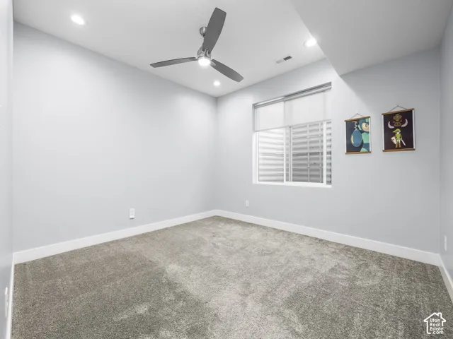 Neutral-toned carpeted room featuring a ceiling fan with integrated lighting, recessed ceiling lights, a window with a roller shade, and white baseboards