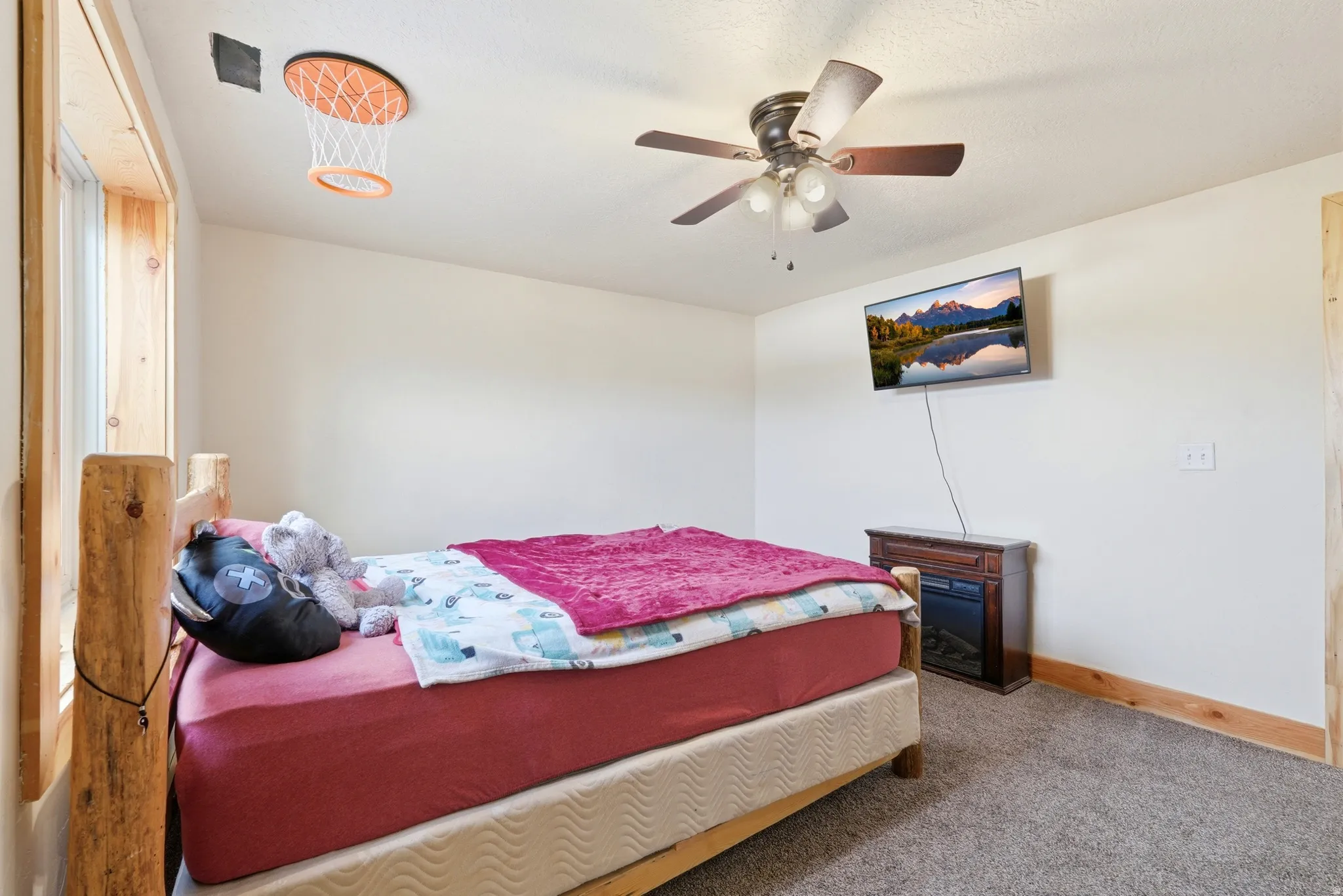 Carpeted bedroom featuring a rustic wood-frame bed, a ceiling fan with integrated lighting, a wall-mounted television, and a built-in electric fireplace