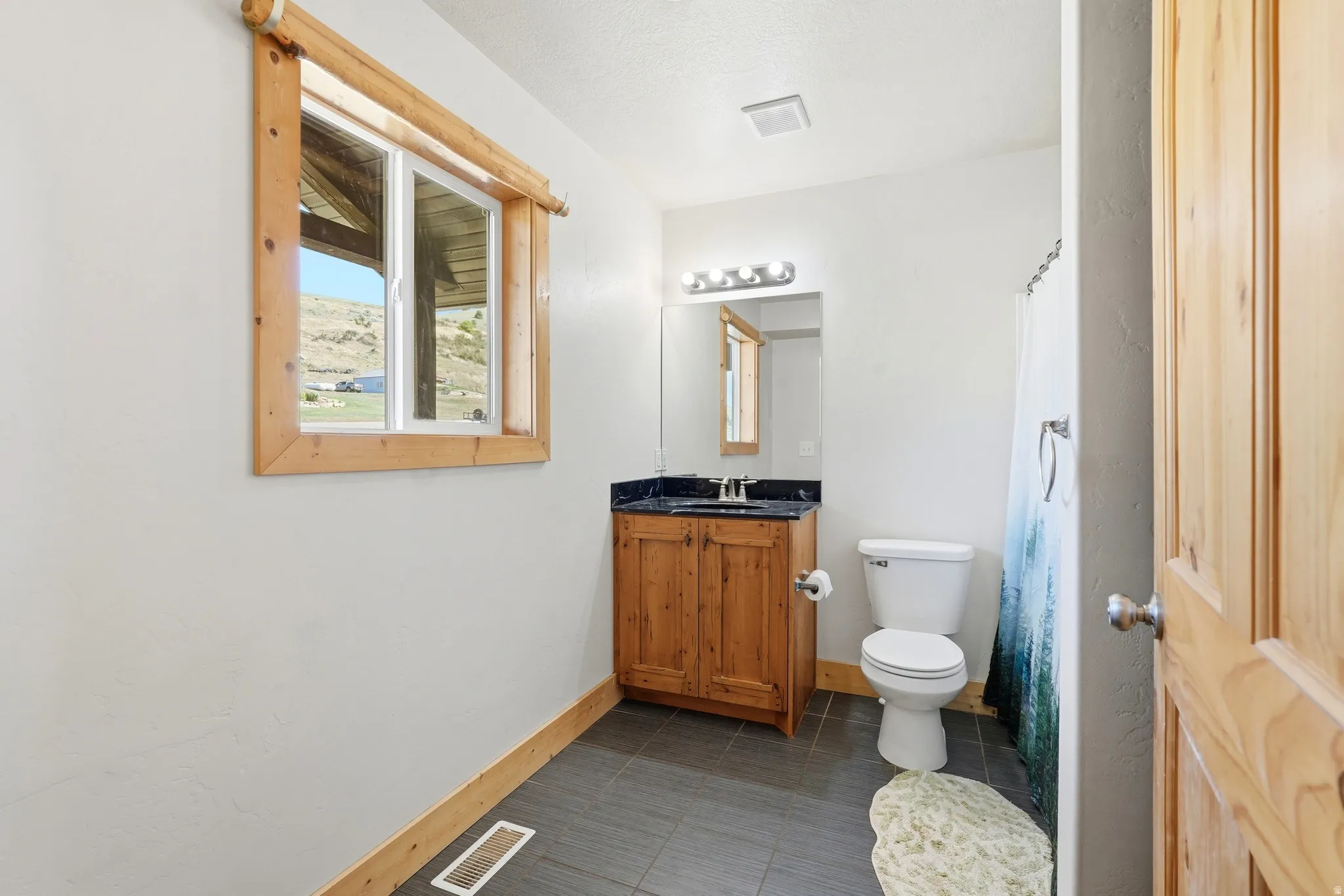 Bathroom featuring a wood-framed window, natural wood vanity with dark countertop, white toilet, and gray tile flooring