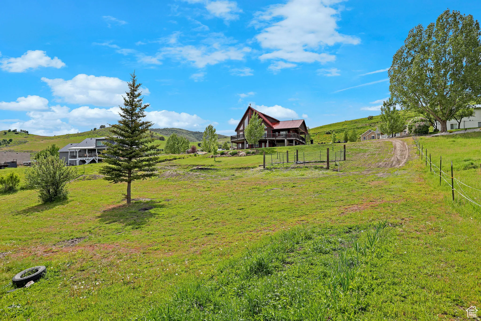 Expansive sloping green landscape with a dirt path leading towards a red-roofed residence featuring a raised deck