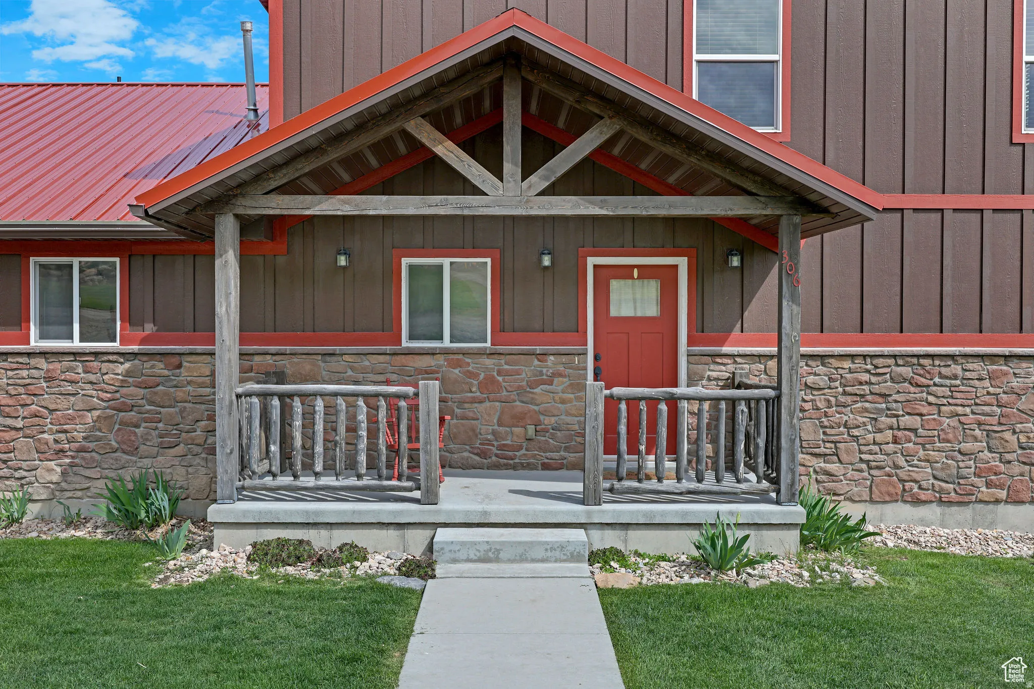 Inviting front entrance featuring a covered porch with log railings, stone facade, board and batten siding, a red metal roof, and a red entry door