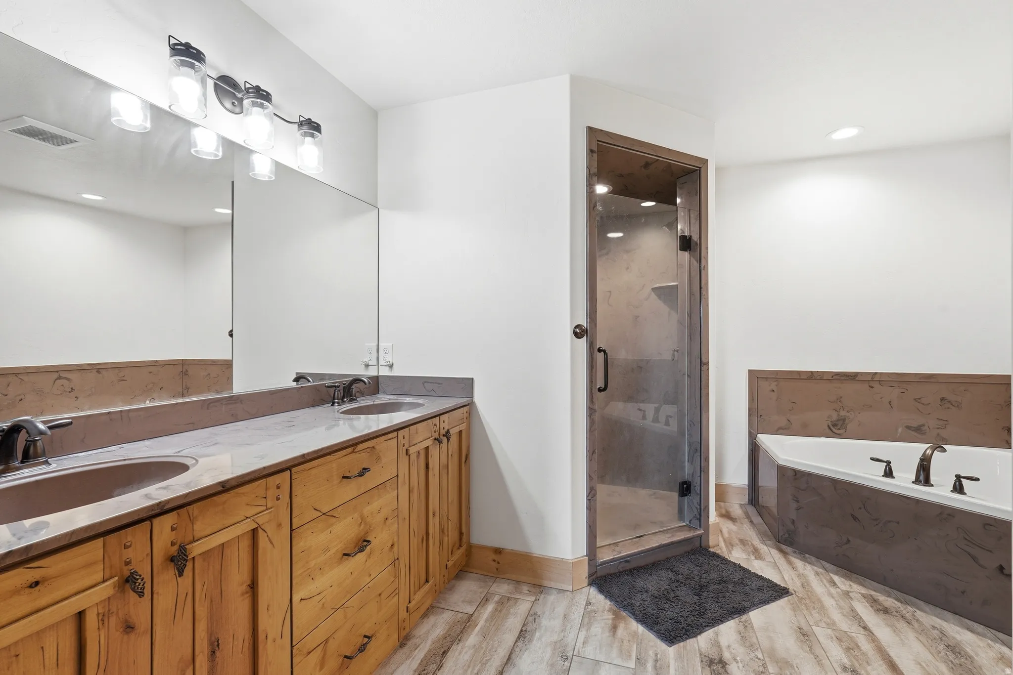Bathroom featuring a double vanity with wood-finish cabinetry and light-toned countertops, a glass-enclosed shower with a wood-trimmed frame, a built-in tub with dark-toned surround, and wood-finish tile flooring