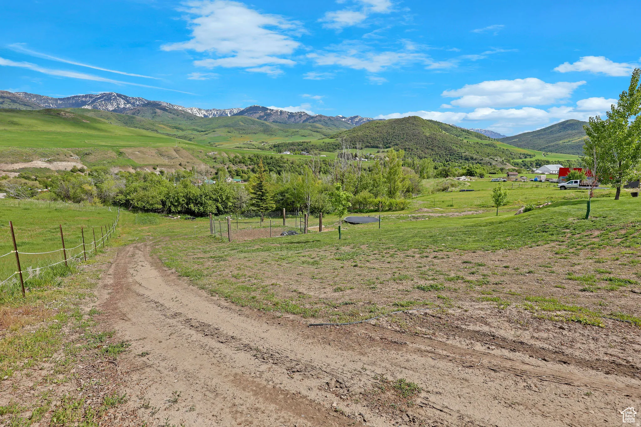 Expansive rural property featuring an unpaved access road, partial fencing, varied topography, mature trees, and distant snow-capped mountains