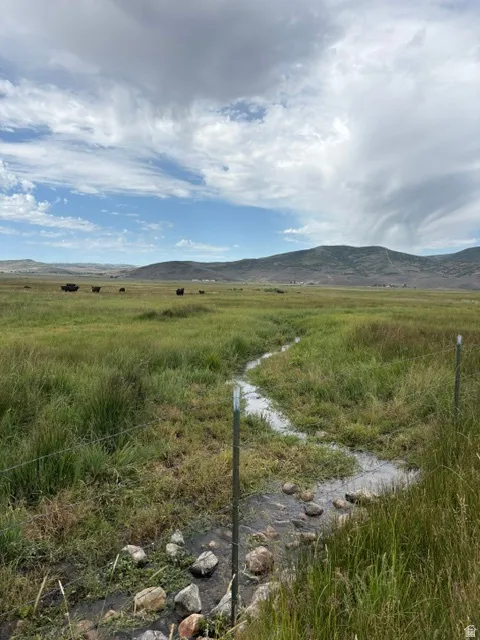 Expansive open pasture with a natural stream, featuring barbed wire fencing and distant mountain ranges under a partly cloudy sky