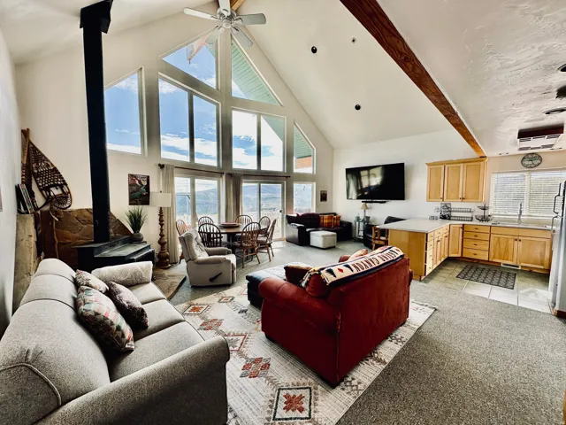 Spacious open-concept living area featuring soaring vaulted ceilings with exposed wood-finish beams, a prominent wood-burning stove with a stone hearth, and a wall of windows