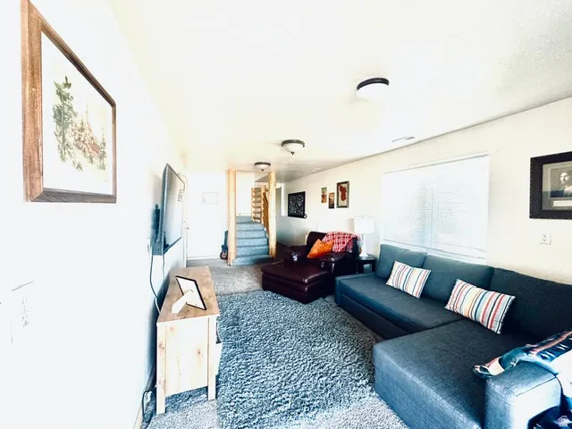Carpeted living area featuring a large window with blinds, ceiling-mounted lighting, and a visible staircase with carpeted treads