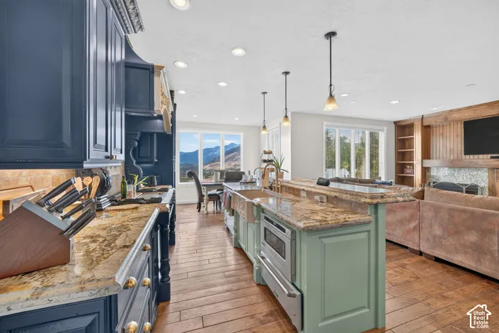 Open-concept kitchen featuring dark cabinetry, natural stone countertops, a light green island with an integrated microwave drawer, and wood-finish flooring