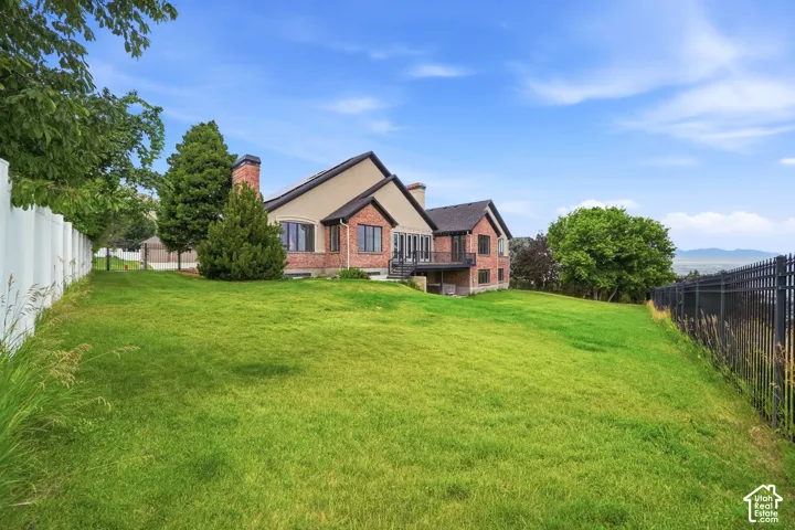 Expansive grassy yard with a multi-level home featuring brick and stucco exterior, multiple windows, an upper-level balcony, and a white privacy fence