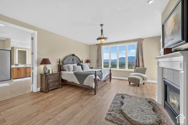 Spacious bedroom featuring wood-finish flooring, a large window with mountain scenery, a white fireplace with a tiled surround, an ornate chandelier, and an adjoining bathroom with a glass-enclosed shower and wood vanity