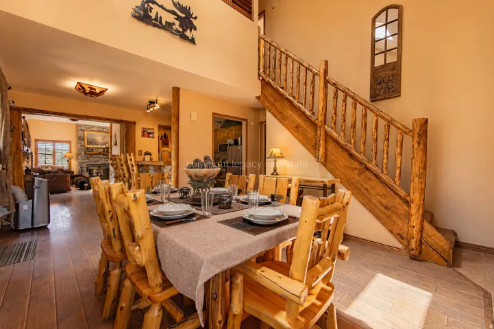 Expansive dining area featuring wood-finish flooring, a rustic wood staircase, and an open entryway to an additional living space with a stone fireplace