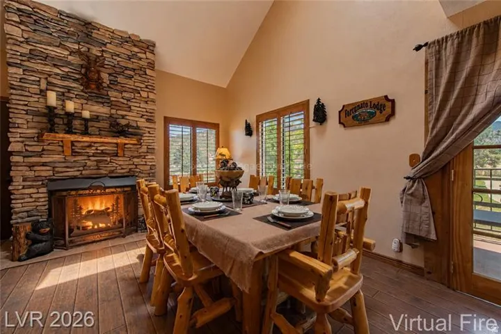 Dining area featuring a stacked stone fireplace, wood-finish flooring, vaulted ceiling, two windows with plantation shutters, and a glass-paneled exterior door