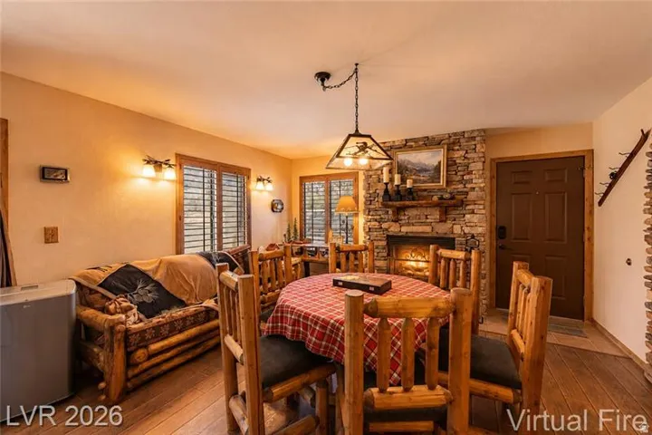 Stone-clad fireplace with mantel and hearth, rich wood-finish flooring, multiple windows with horizontal blinds, built-in wall sconces, and a paneled entry door