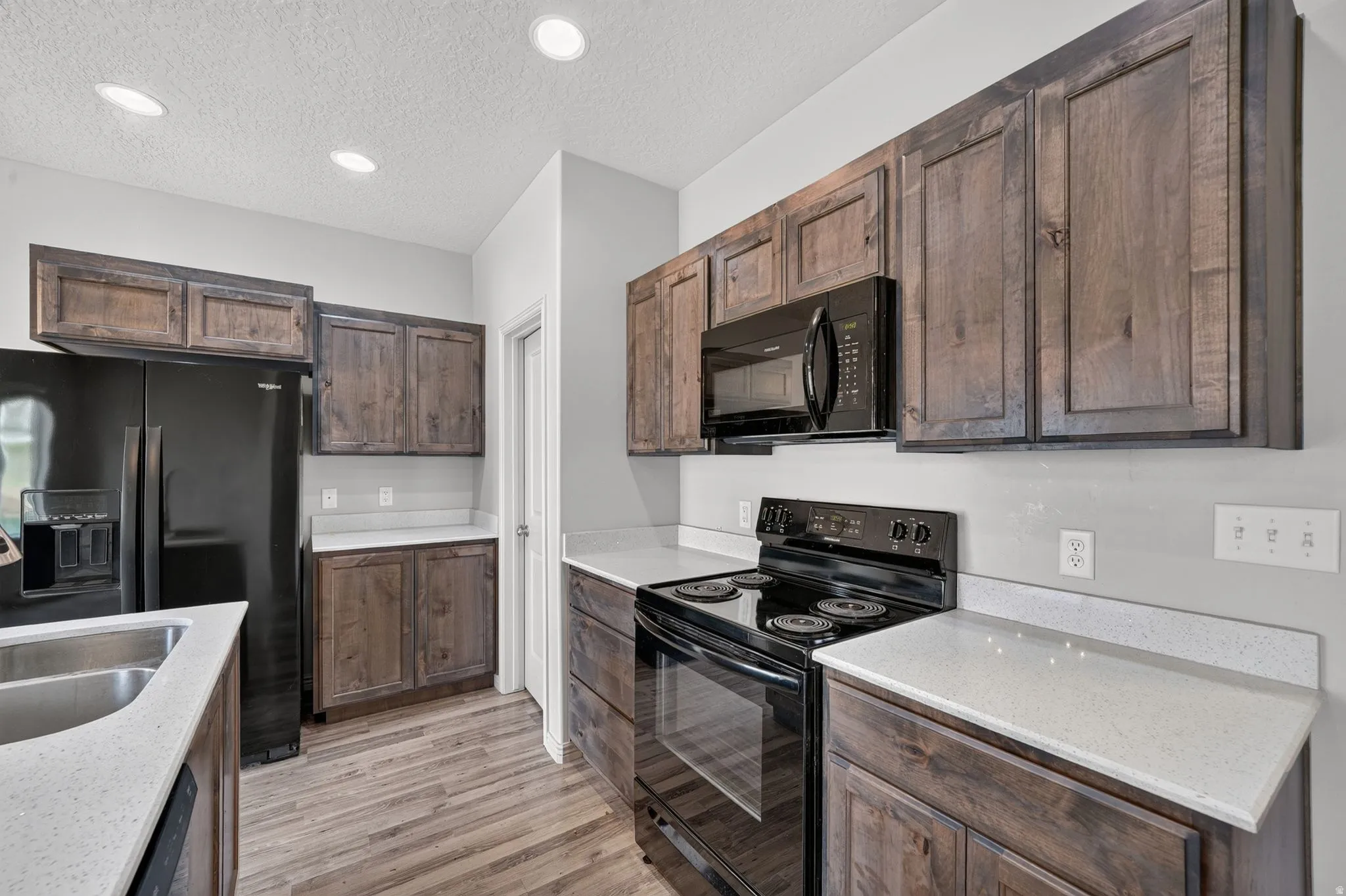 Kitchen featuring dark wood-finish cabinetry, light-toned countertops, wood-finish flooring, a double basin stainless steel sink, and black appliances