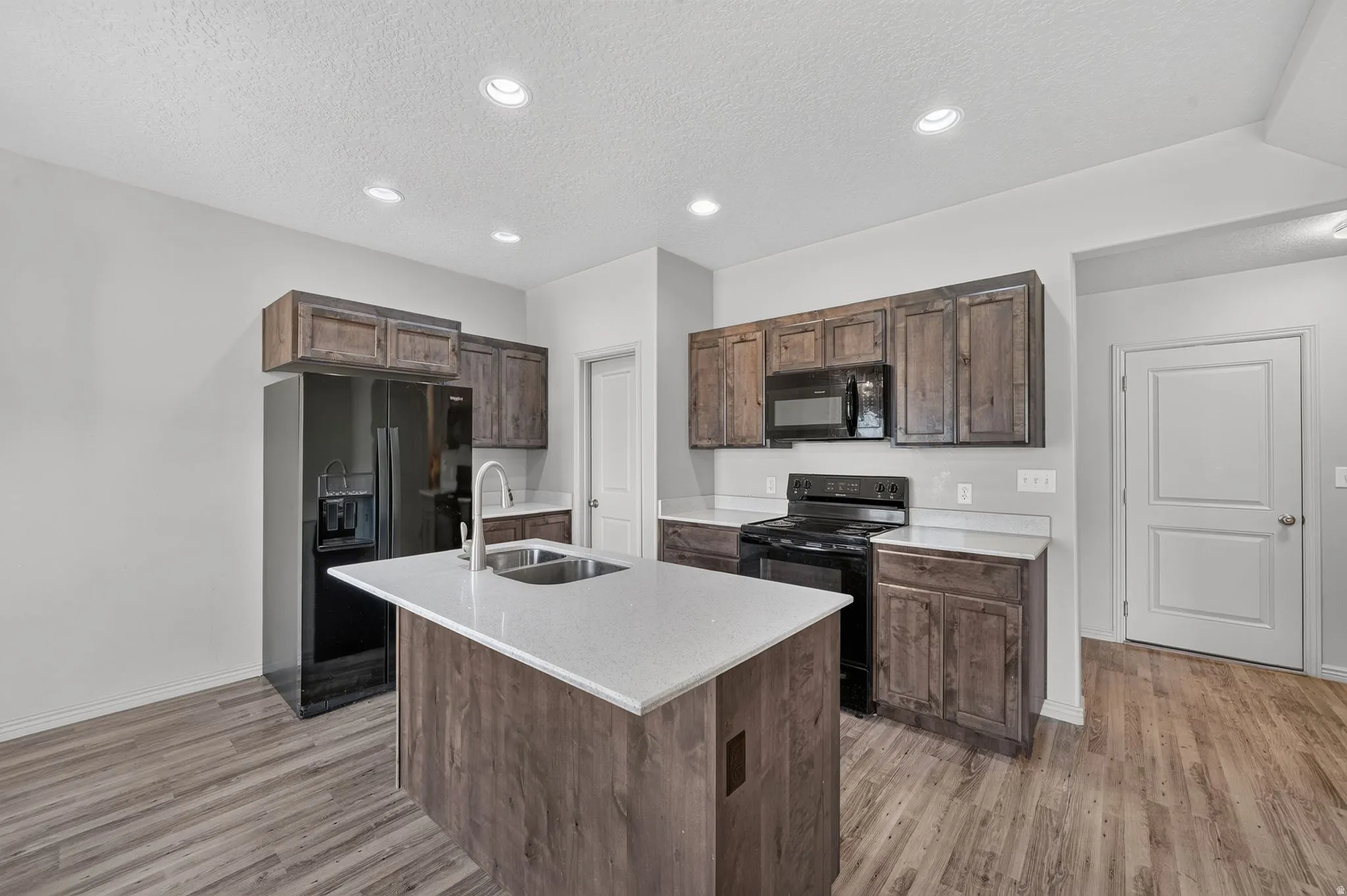 Kitchen featuring a central island with a double basin sink, white countertops, dark wood-finish cabinetry, black appliances, and recessed ceiling lighting