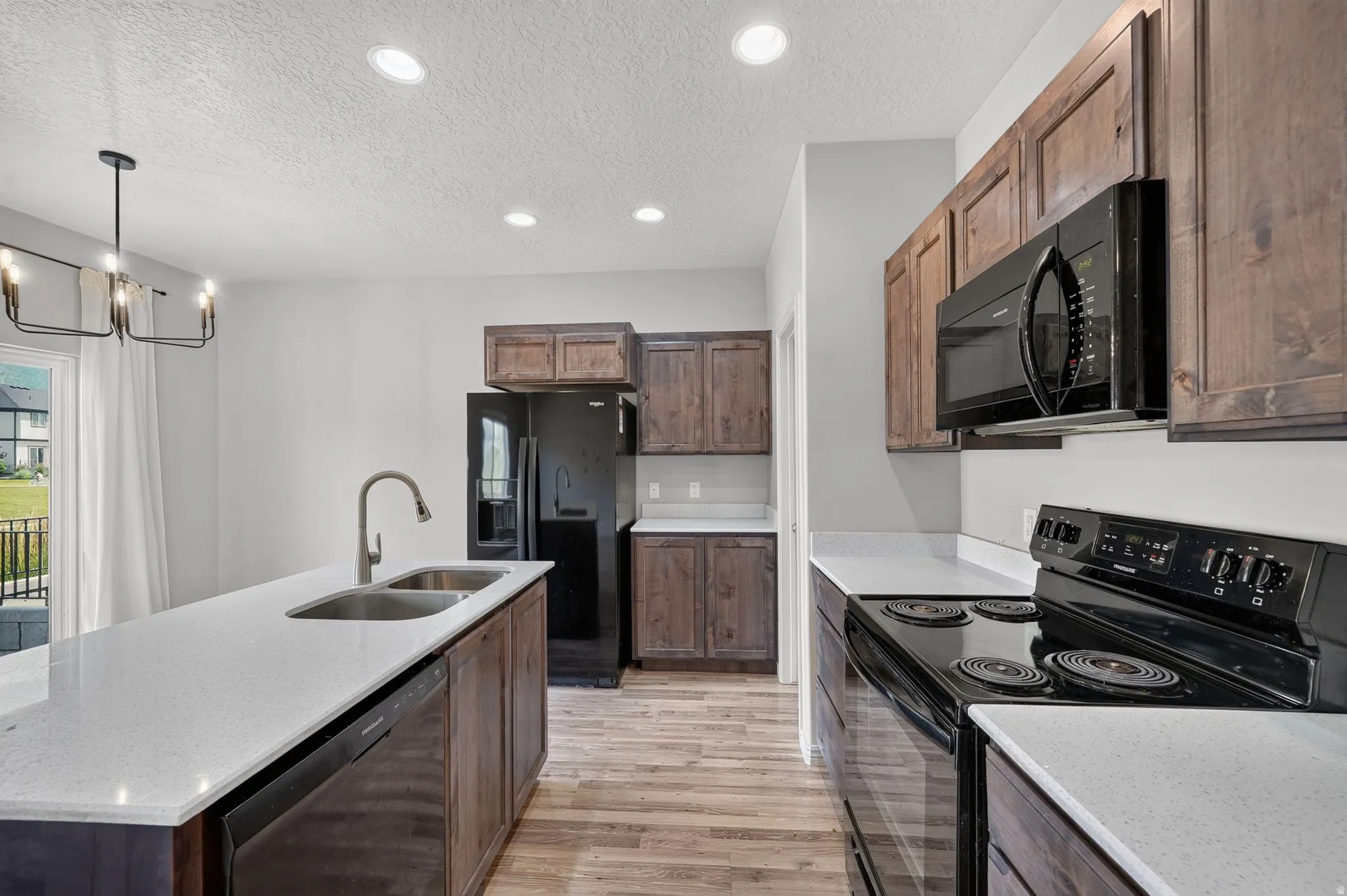 Modern kitchen featuring a large island with an undermount double-basin sink, light-toned countertops, dark wood-finish cabinetry, and black appliances