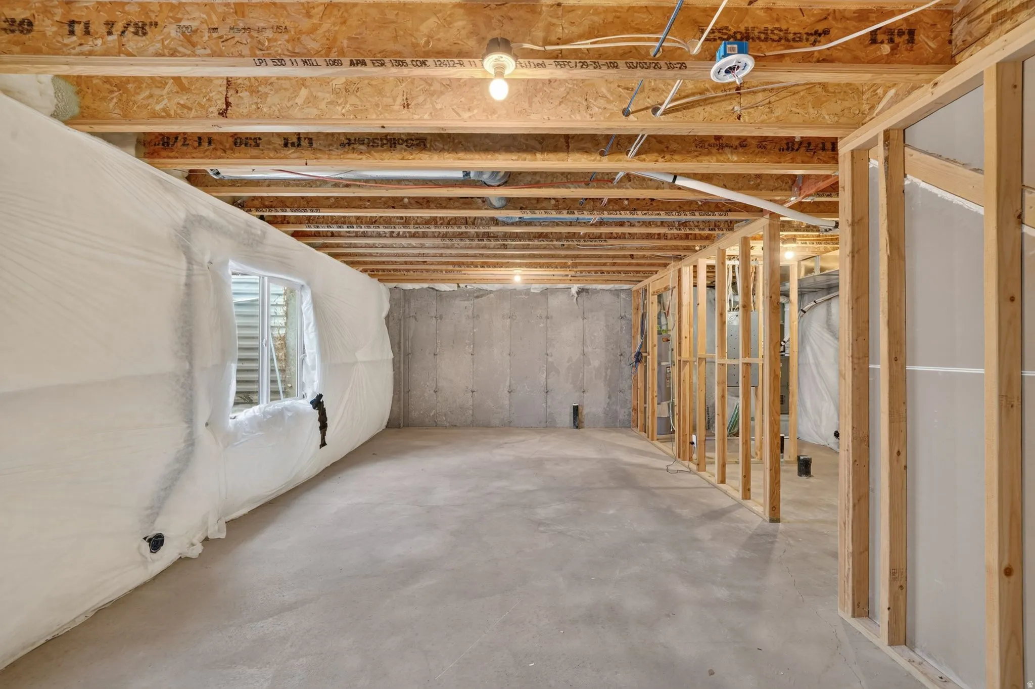 Unfinished basement featuring exposed wood joists, concrete flooring, framed interior walls, insulated exterior wall, and an egress window