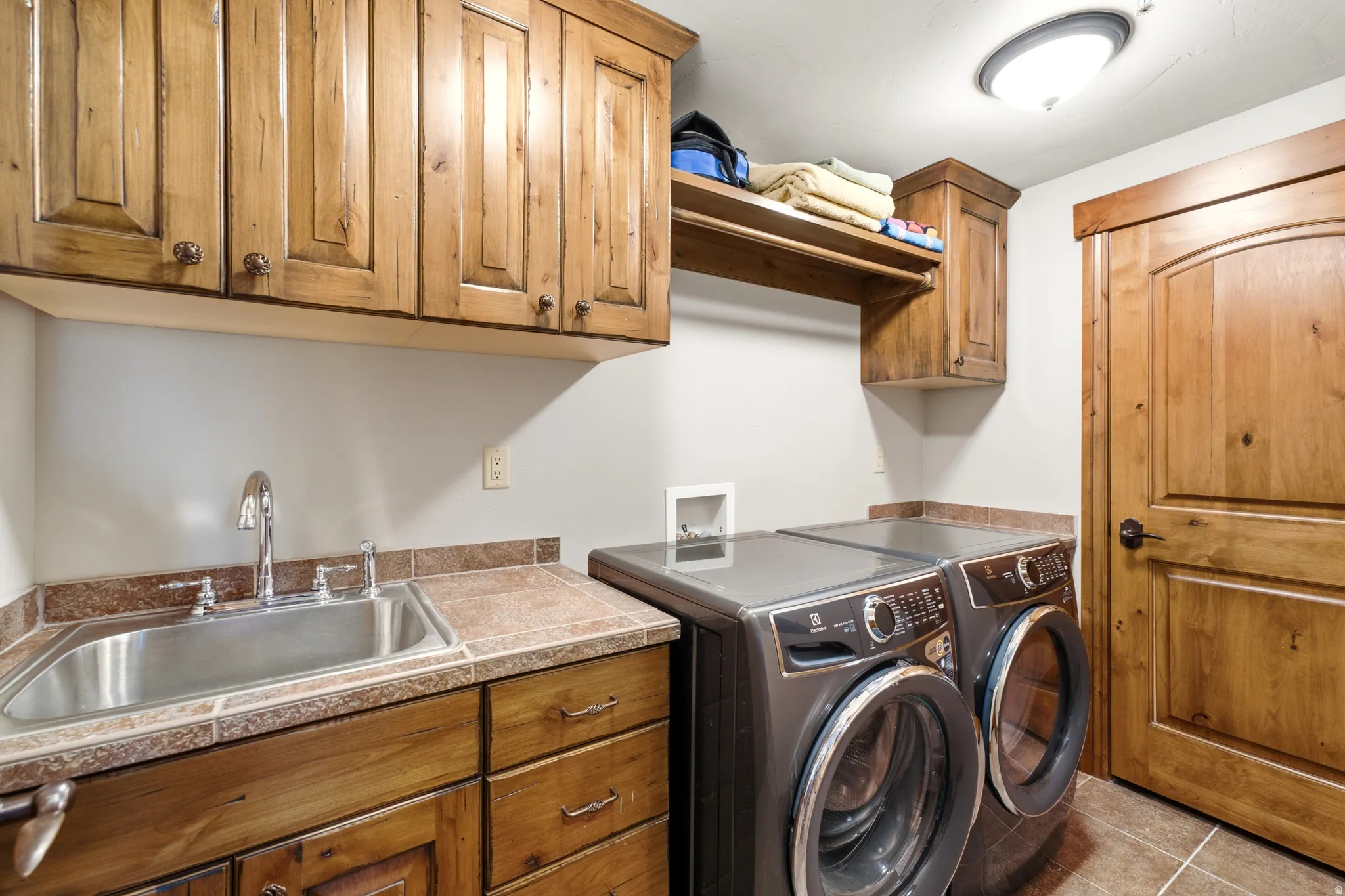 Laundry room featuring wood-finish cabinetry, a utility sink with a stainless steel basin, and tiled countertops