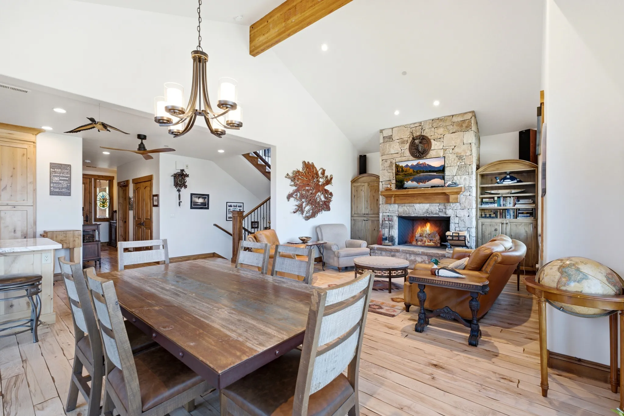 Spacious open-concept living area featuring high vaulted ceilings with exposed wood beams, a prominent stone fireplace with a substantial wood mantel, a multi-light chandelier, and light wood-finish flooring