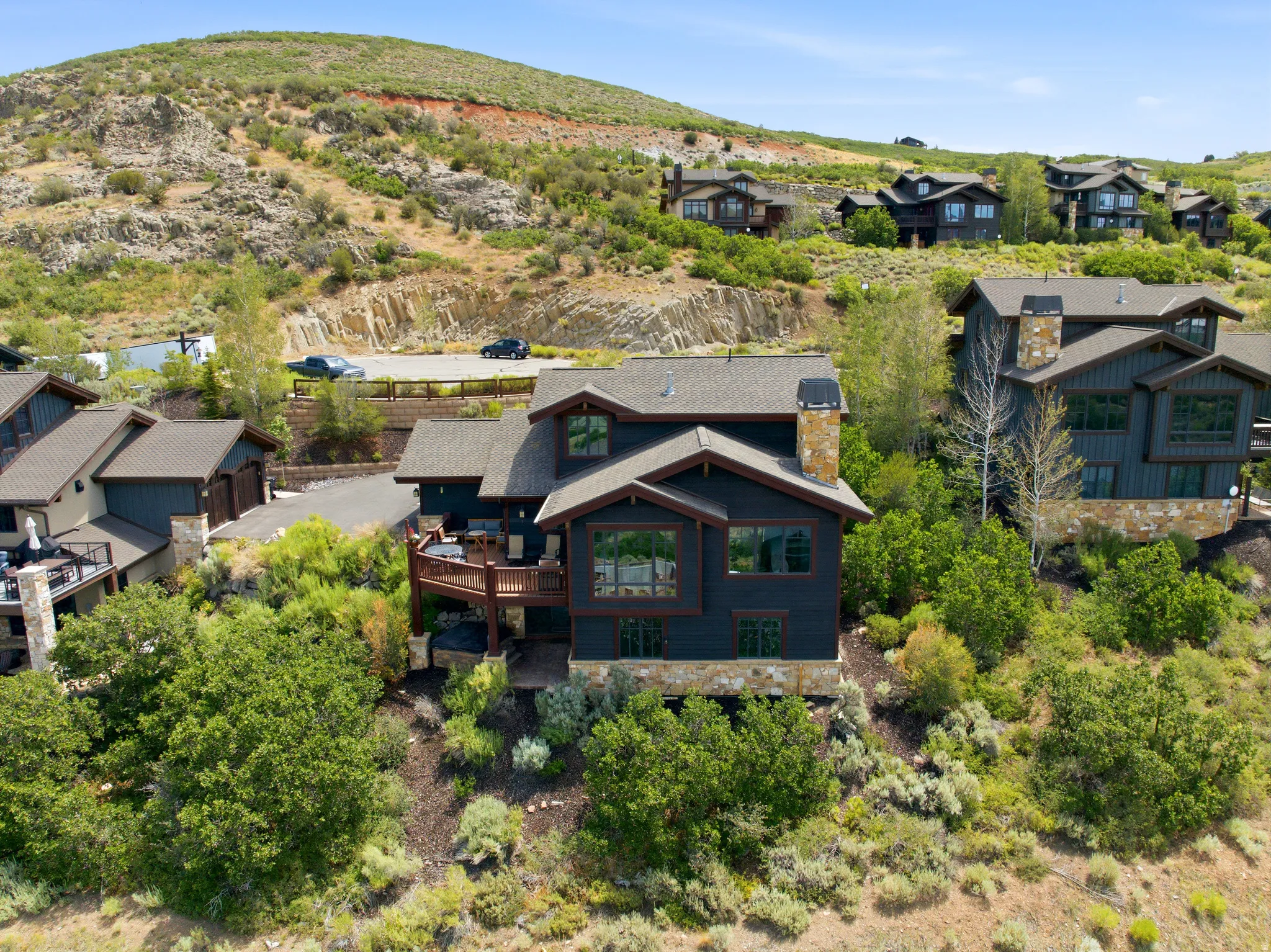 Modern mountain architecture featuring a dark exterior, stone accents, multiple large windows, a spacious wood deck, and a stone chimney