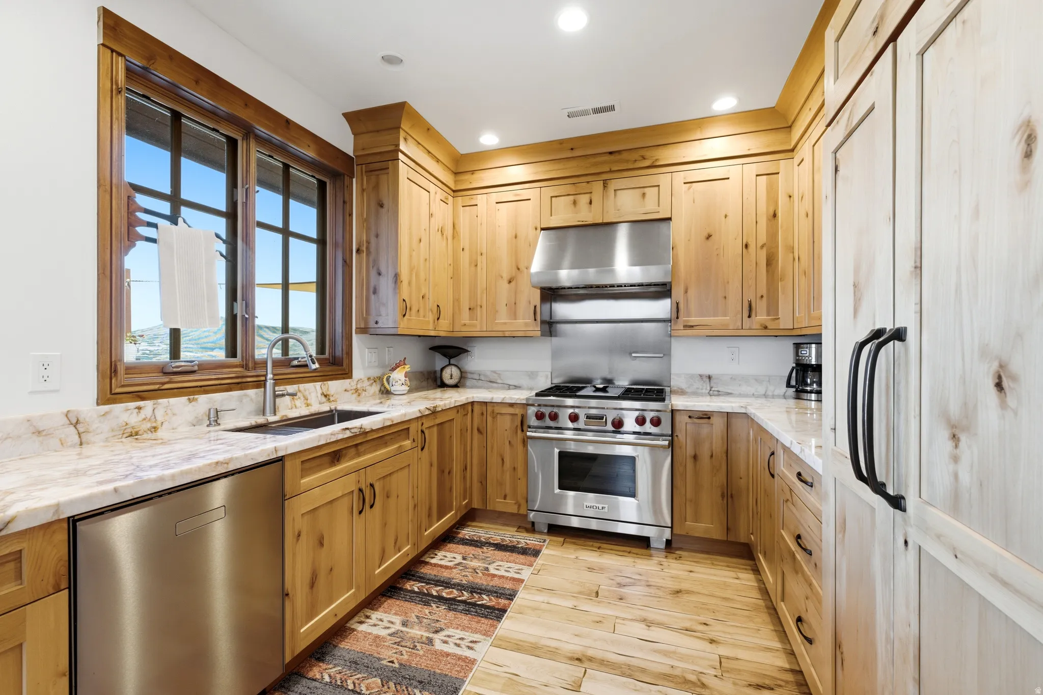 Kitchen featuring extensive natural wood cabinetry, light-toned stone countertops, and a full suite of stainless steel appliances including a range with hood and a paneled refrigerator