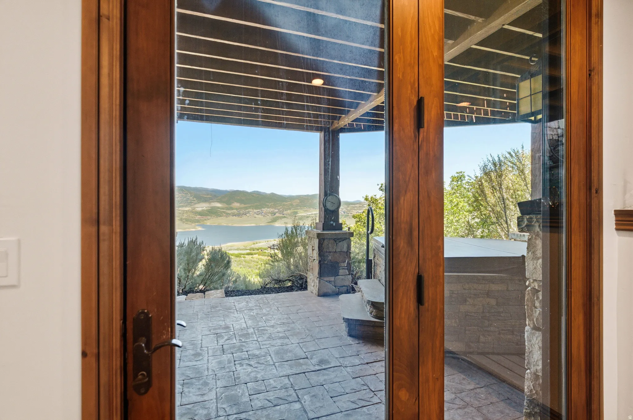 Wood-framed glass doors open to a stamped concrete patio featuring a built-in hot tub, stone-clad pillars, and a mountainous lake backdrop