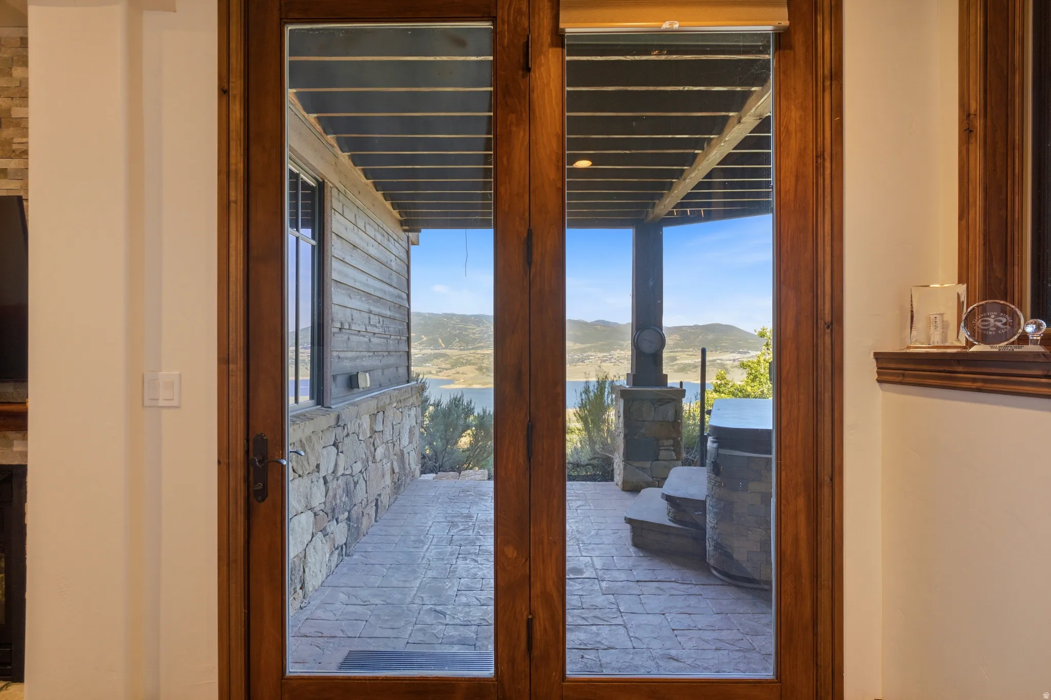 Double glass-paneled doors with wood-finish frames open to an outdoor patio featuring stamped concrete flooring, a built-in hot tub with stone surround, and stone pillars