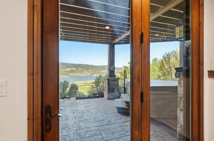 Wood-framed glass doors open to a stamped concrete patio featuring a built-in hot tub, stone-clad pillars, and a mountainous lake backdrop