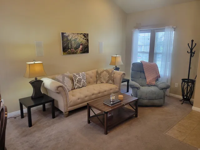 Living area featuring a soft beige color palette, light tan carpet flooring, and a large window with privacy blinds