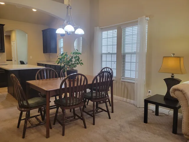 Open-concept area featuring a dining space with an overhead chandelier, two large windows with blinds, a kitchen island with a light-colored countertop, dark cabinetry, and light beige wall paint