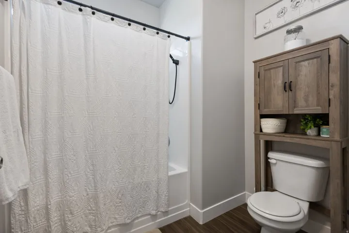 Tub and shower combination featuring a black shower head and rod, complemented by a wood-finish over-toilet storage cabinet with matte black hardware