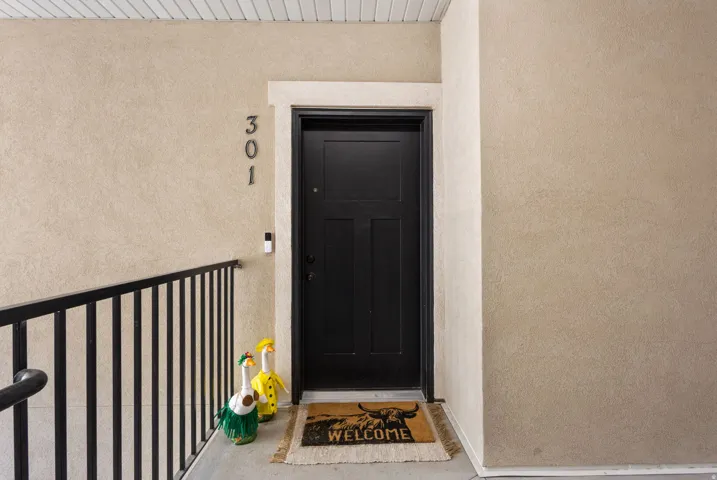 Entryway featuring a black paneled door, textured exterior wall finish, black metal railing, and white ceiling soffit