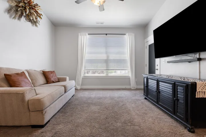 Carpeted interior room featuring light gray wall paint, a window with horizontal blinds and white curtains, and a ceiling fan with integrated lighting