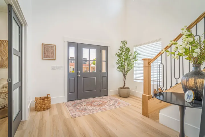 Entryway featuring light wood-finish flooring, a contemporary dark gray front door with glass panels, a white accent wall, a staircase with a natural wood handrail and black iron balusters, and a side window with horizontal blinds