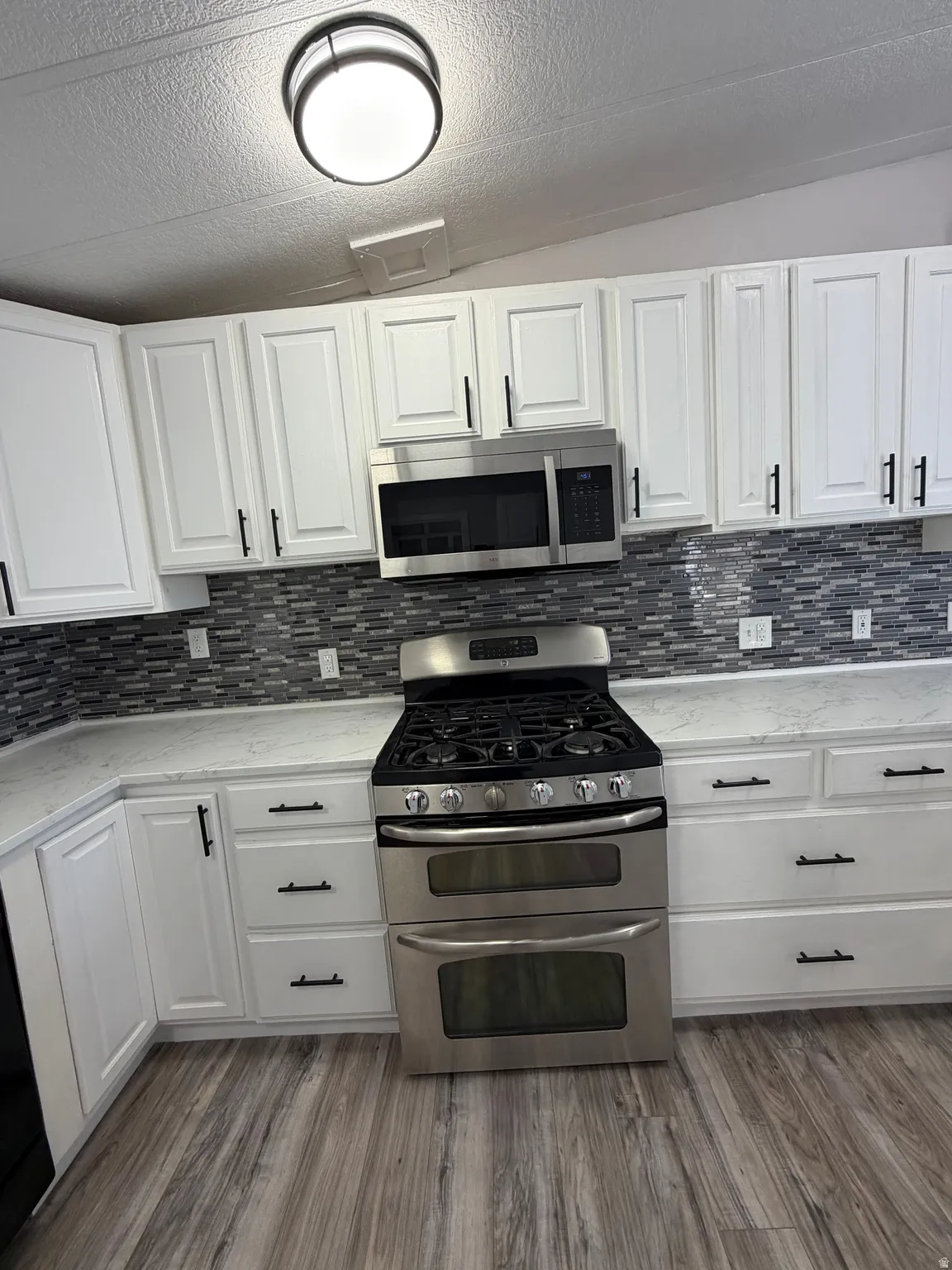 Contemporary kitchen featuring white cabinetry with matte black hardware, stainless steel appliances including a double oven range and microwave, light-toned countertops, and a gray mosaic tile backsplash